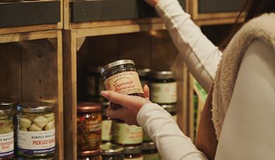 A picture of a lady browsing the shelves of a shop, holding a jar in her left hand and reaching for a bottle of red wine with her right hand.