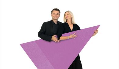 Two musicians are stood smiling and holding a purple triangle graphic.