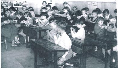 Black and white photograph of a school classroom. Children can be seen sitting at their desks working together.