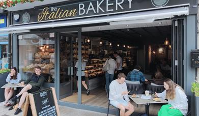 Front of The Italian Bakery with people sat at outdoor tables enjoying coffee and cakes. Open front to the shop exposing the interior counters filled