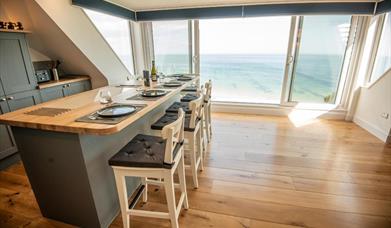 Modern kitchen island counter on hardwood flooring in front of a wall sized window with a glass Juliet balcony overlooking the sea