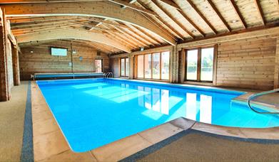 Indoor heated swimming pool and sauna inside a wooden lodge with floor to ceiling windows and the sunshine pouring through the windows.